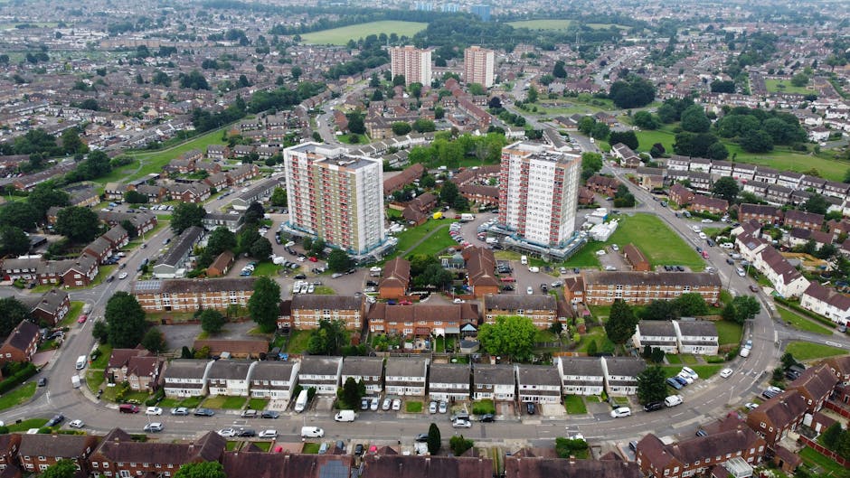 Aerial view of a residential neighbourhood showing rows of terraced houses with driveways and parked cars in the foreground. Behind them, there are two high-rise apartment buildings with distinct whit