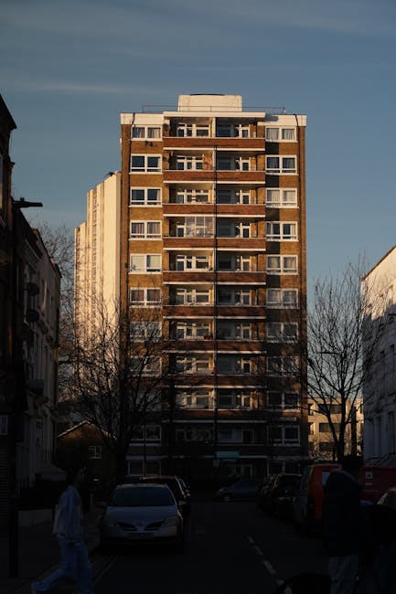 Aerial view of a residential neighbourhood showing rows of terraced houses with driveways and parked cars in the foreground. Behind them, there are two high-rise apartment buildings with distinct whit
